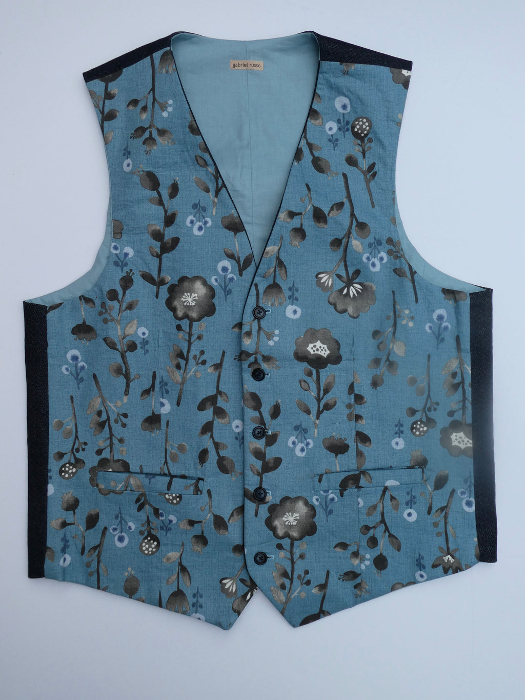 A sleeveless vest with a dusty blue base and an all-over floral pattern in brown, gray, and white. It features black side panels and black trim along the neckline and armholes, photographed flat against a light gray background.