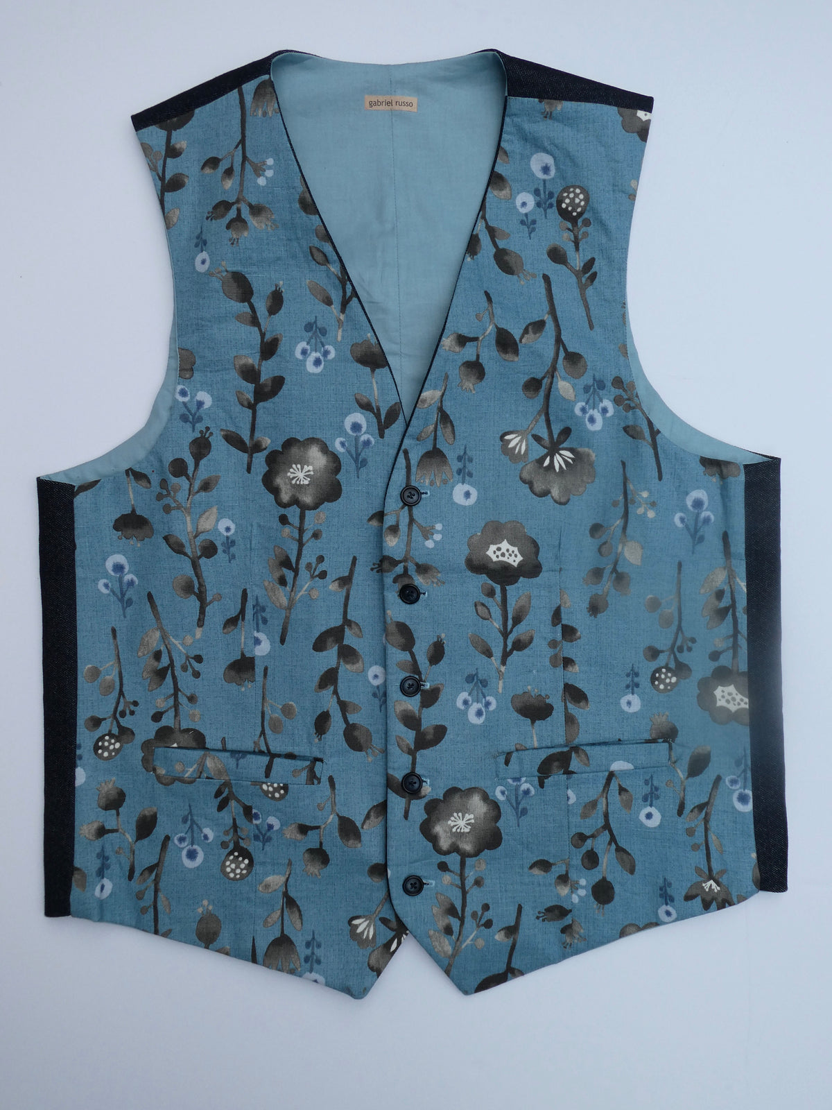 A sleeveless vest with a dusty blue base and an all-over floral pattern in brown, gray, and white. It features black side panels and black trim along the neckline and armholes, photographed flat against a light gray background.